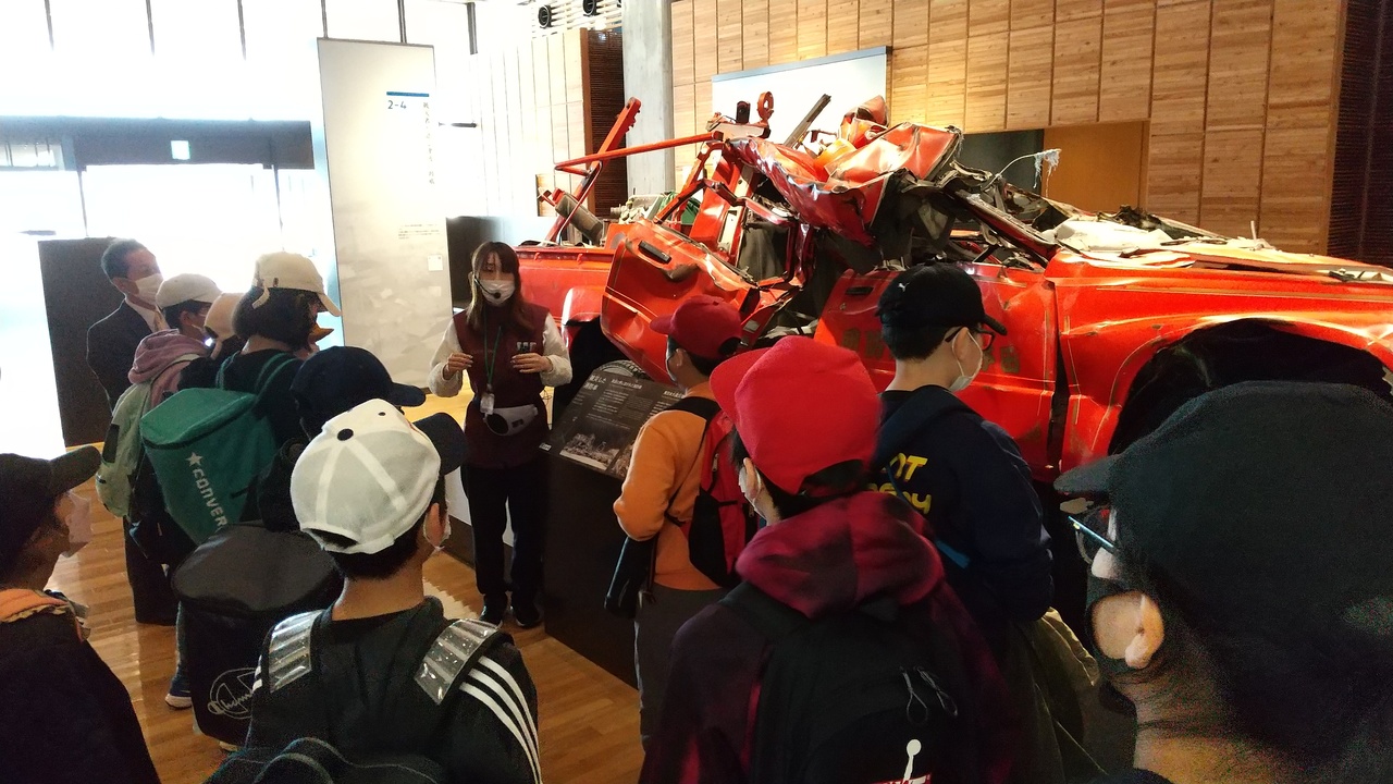 展示されている破損した赤い車の前に立つインターカムをつけた女性と、その周囲に集まる子供たちの写真