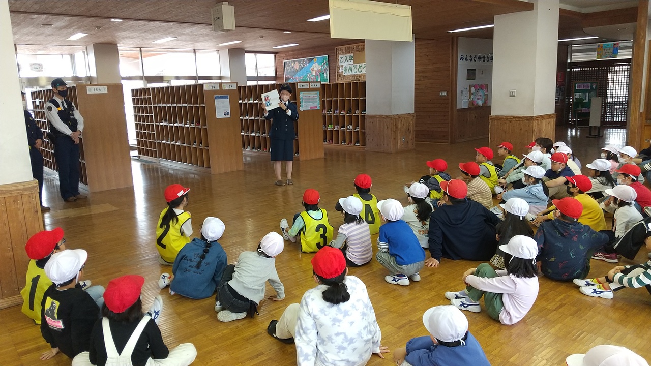 下駄箱がある校舎の出入口の前で児童たちが体育帽子を被って座り、婦警の話を聞いている写真