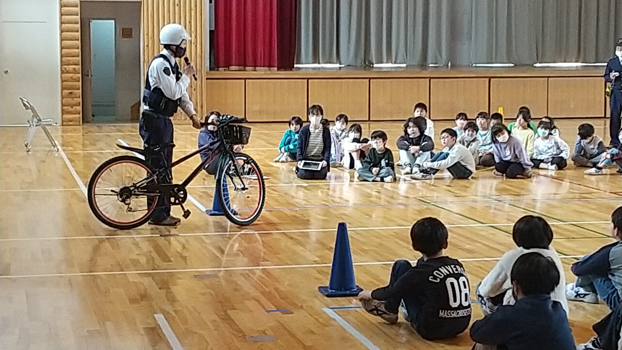 体育館で児童たちが座っており自転車を脇に置いている警官から話を聞いている写真