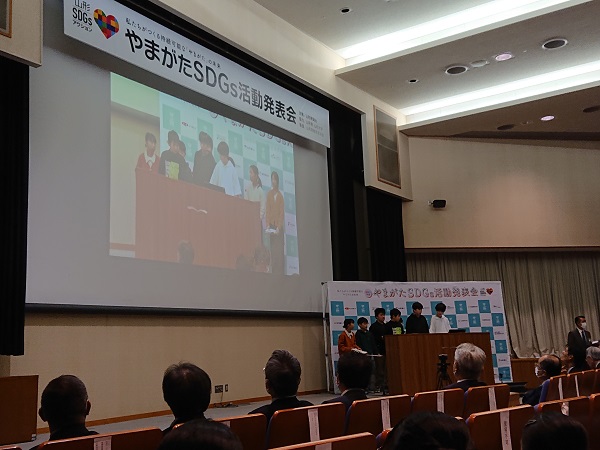 「やまがたSDGs活動発表会」と書かれた大きなスクリーンの下で、子供たちが壇上で横一列に並んで発表している写真