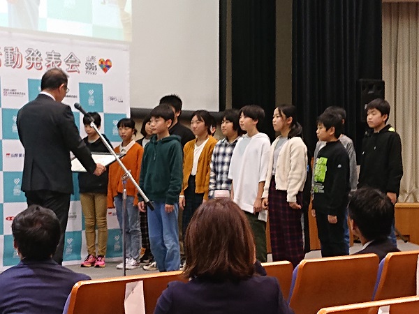 スーツ姿の男性が賞状を持って子供たちに向き合っている写真