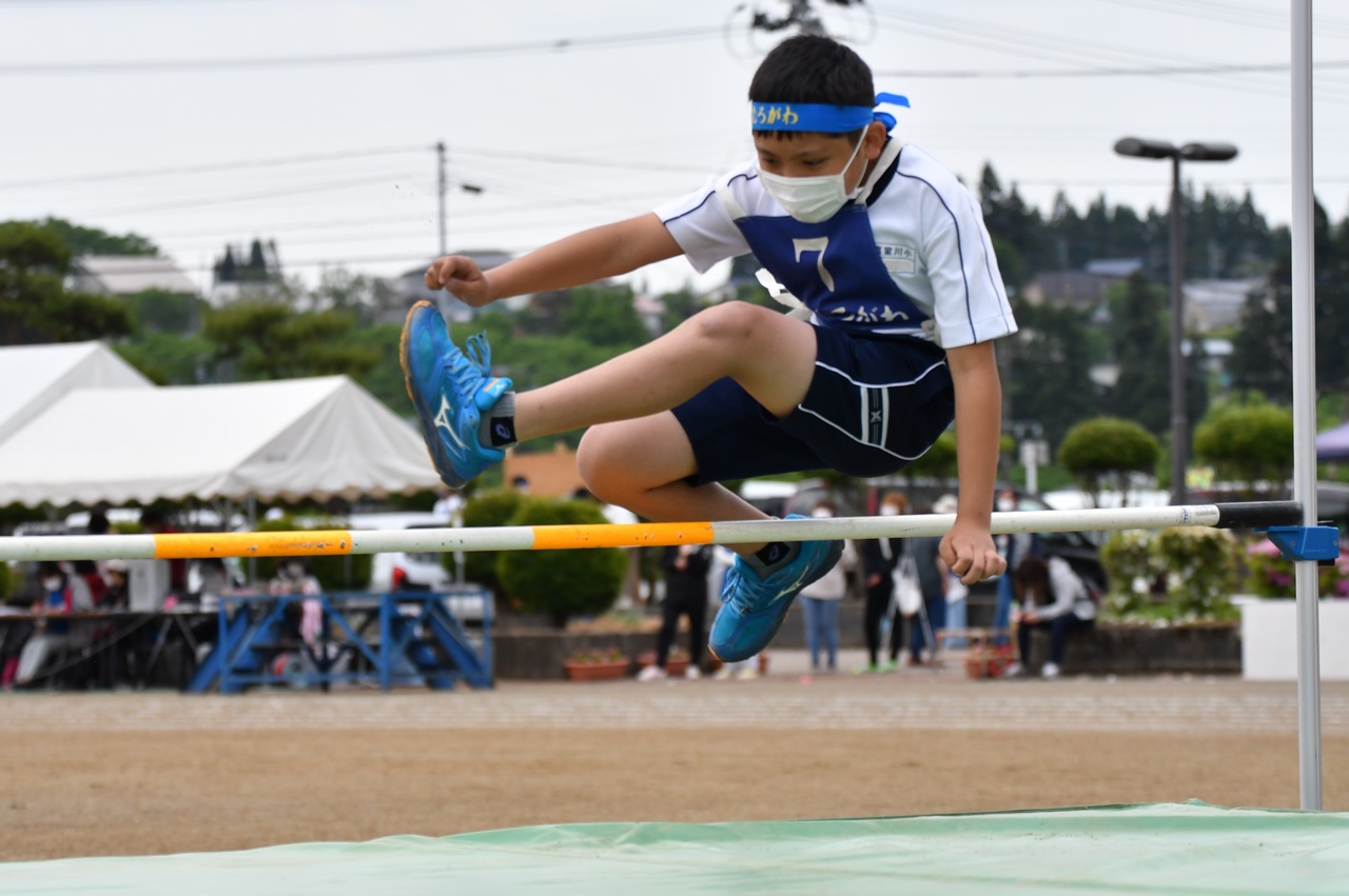 青い鉢巻に体操服姿の男の子が走り幅跳びのバーを越えている写真