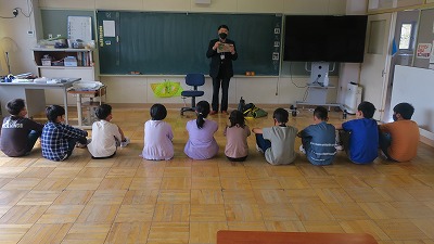 床に一列に並んで座り、読み聞かせをして下さる男性の声に聴き入る児童たちの写真