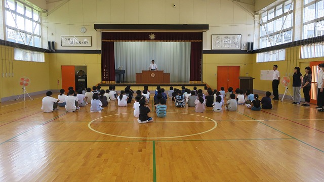体育館の床に座り校長先生の話に耳を傾ける児童たちの写真
