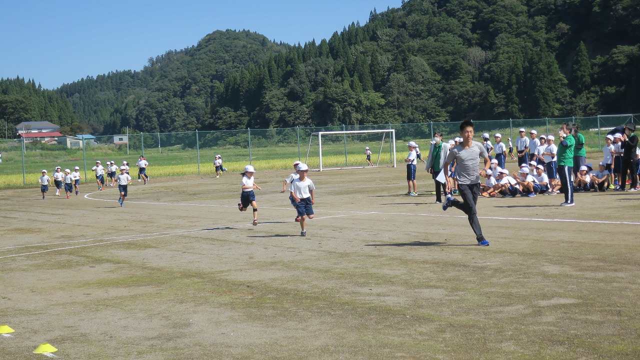小学校の運動場で前方を走っている駅伝選手に追いつこうと頑張る低学年の児童たちの写真