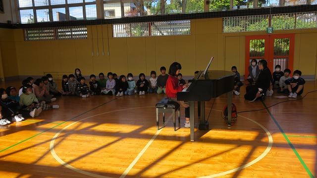 児童と先生の前でピアノ演奏を披露する赤い服を着た女子児童の写真