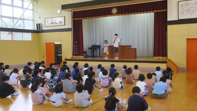 壇上にに上った2人の児童代表の話を熱心に聴く児童たちの写真