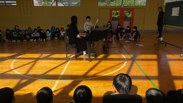 児童と先生たちの前でピアノの伴奏に合わせて歌を披露する男子児童の写真