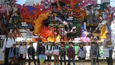 校外学習にて、新庄まつりの「山車」を背景に撮影された、教職員の男女2人と、2年生の児童11人の集合写真