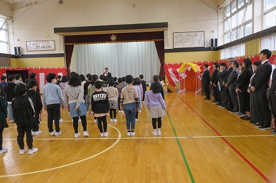 小学校の体育館で行われた1学期の始業式にて、壇上前で話をするスーツを着た男性と、その話を立ちながら聞いている大勢の児童達と、その右隣に立つ教師陣の様子を写した写真