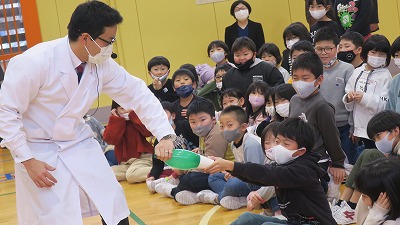 白衣を着た大人の男性と男子児童が、何か緑の物を互いに引っ張り合っている写真