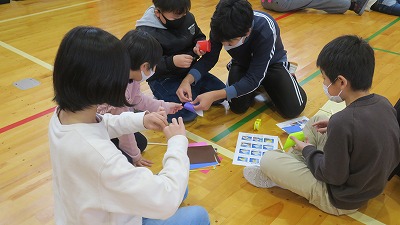 体育館の床に座って、児童たちが小さな絵のようなものをやり取りしている写真
