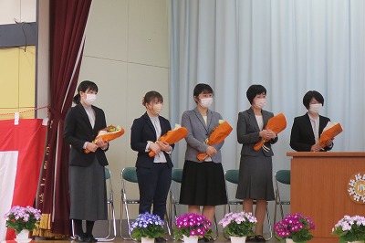 小学校の体育館で行われた離任式にて、壇上で花束を持って並んで立っている、離任予定の5人の女性教諭の様子を写した写真
