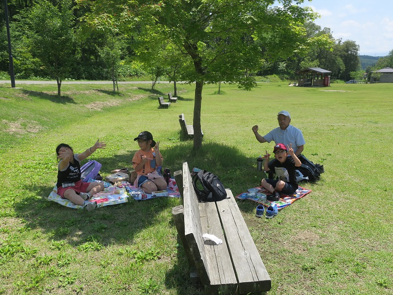 日を避けるように芝生の木陰になった部分にシートを敷いてお弁当を食べる先生と児童たちの写真