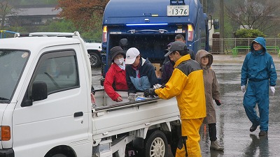 雨のなか、青のゴミ収取車から白の小型トラックの荷台に、回収した新聞紙・雑誌・アルミ缶・段ボールを載せ替えている複数人の高齢の男女の様子を写した写真