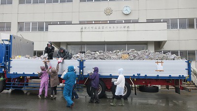 雨のなか、大型トラックの荷台に、回収した新聞紙・雑誌・アルミ缶・段ボールを載せている複数人の高齢の男女の様子を写した写真