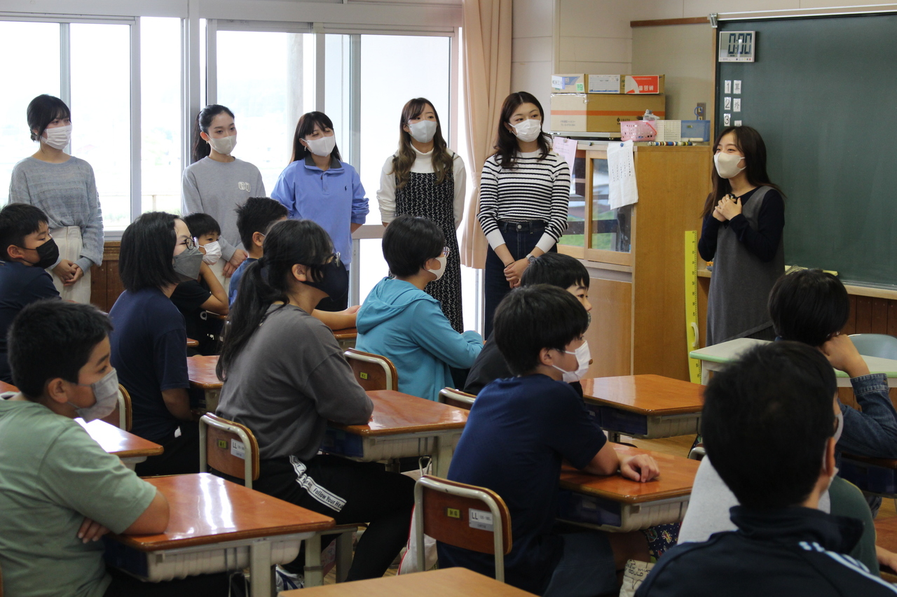 黒板の前で話す女性を窓際に立つ女性5人と席についた子供たちが眺めている写真