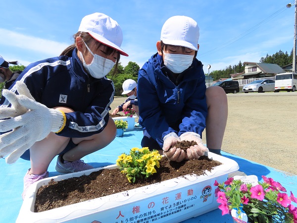 ジャージ姿の子供2人がプランターに黄色い花の苗を植えている写真