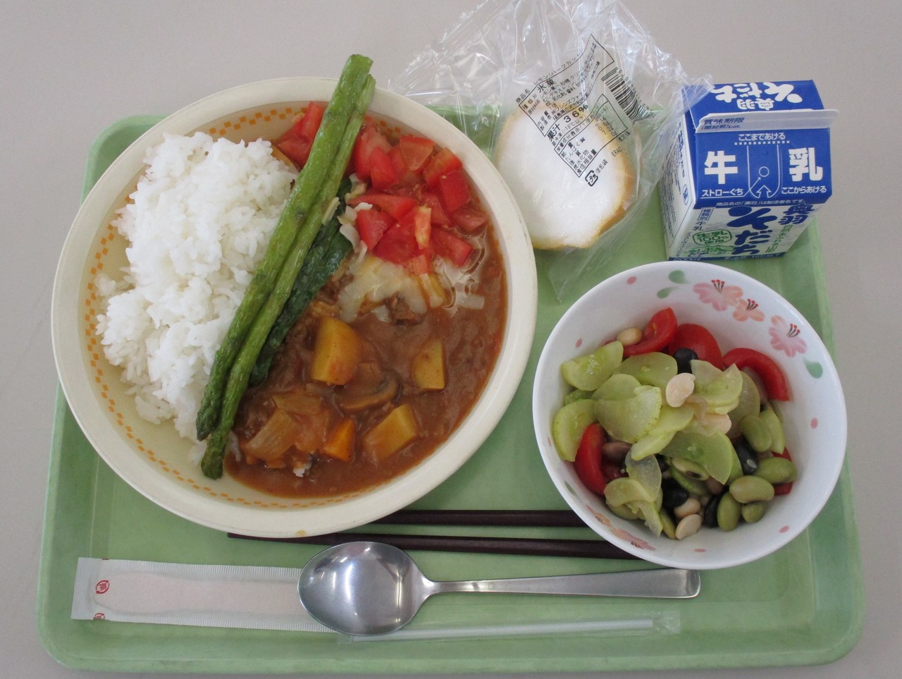 丸ごとのアスパラガスが上に乗っているカレーと副菜などの並んだ給食の写真