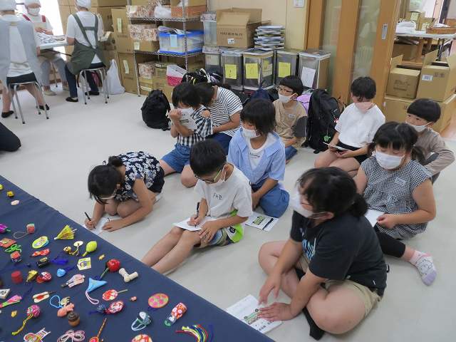 宇佐美煎餅店で扱っているおもちゃを見てメモに書き留めている児童たちの写真