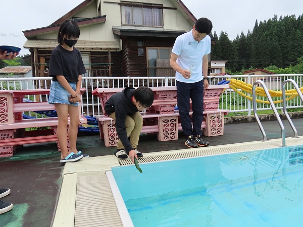 緑の棒をプールにつけている男の子と、その様子を見守る女の子と男性の写真