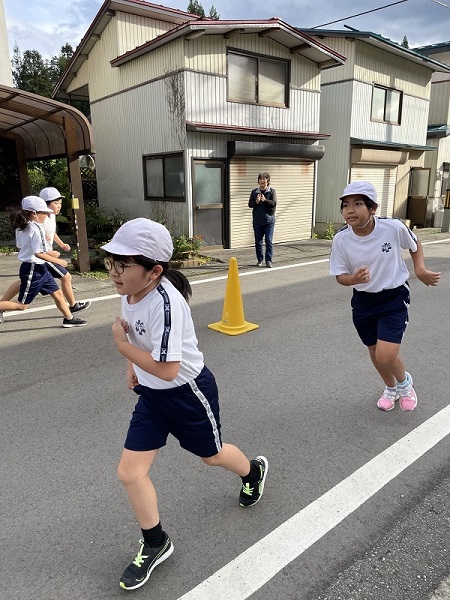 黄色いコーンの置かれた民家前の道路を体操服姿の子供たちが走っている写真