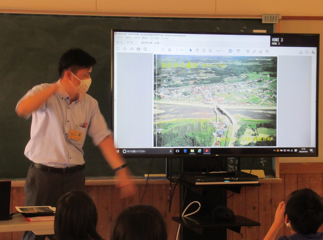 座った小学生の前で胸元にカードを付けた男性と航空写真が映し出されたテレビの写真