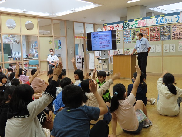 モニターの横の講演台に立つスーツ姿の男性と、床に座って手を挙げる子供たちの写真