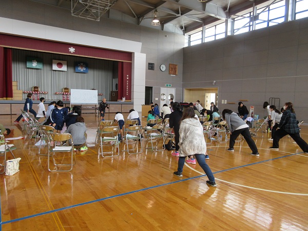 体育館でパイプ椅子に座っている人たちとその手前でアキレス腱を伸ばす運動をしている女性たちの写真