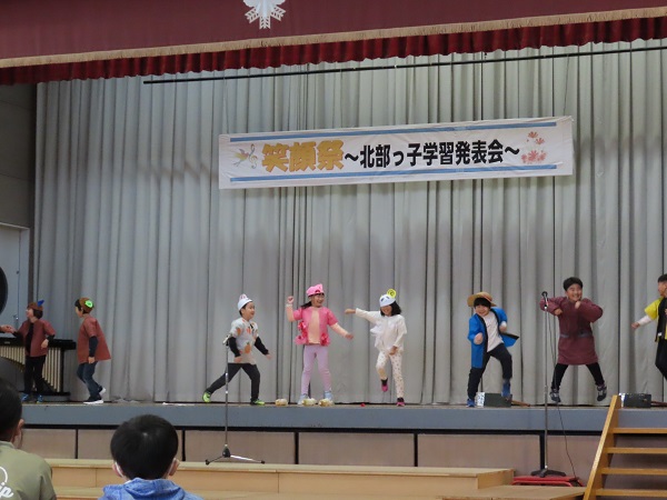「笑顔祭北部っ子学習発表会」と書かれた垂れ幕の下、壇上で劇を披露する子供たちの写真