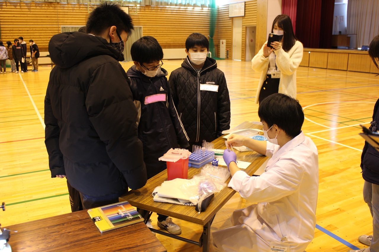 白衣を着て試験管を扱っている男性の作業を見ている男子児童たちの写真