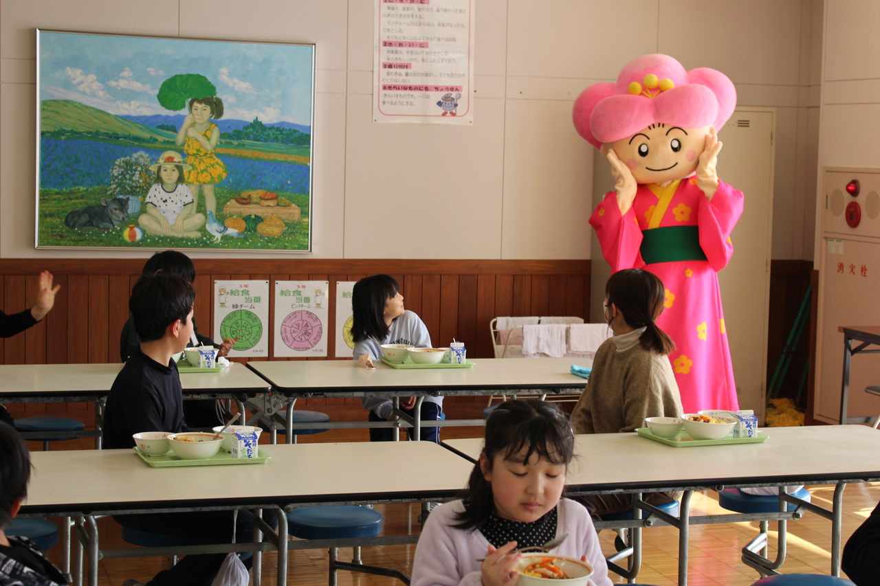 席について給食を食べる子供たちがピンク色の和服を着た着ぐるみを眺めている写真