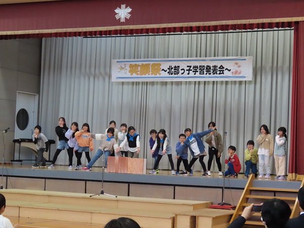 「笑顔祭北部っ子学習発表会」と書かれた垂れ幕の下、壇上で発表をする大勢の子供たちの写真
