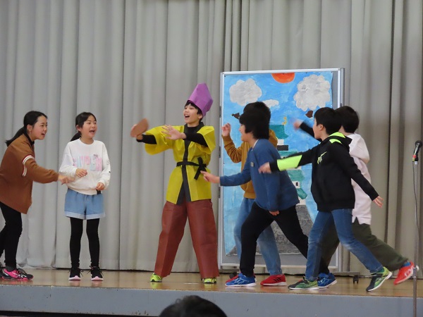 青空と城のイラストの前で、劇を演じる子供たちの写真