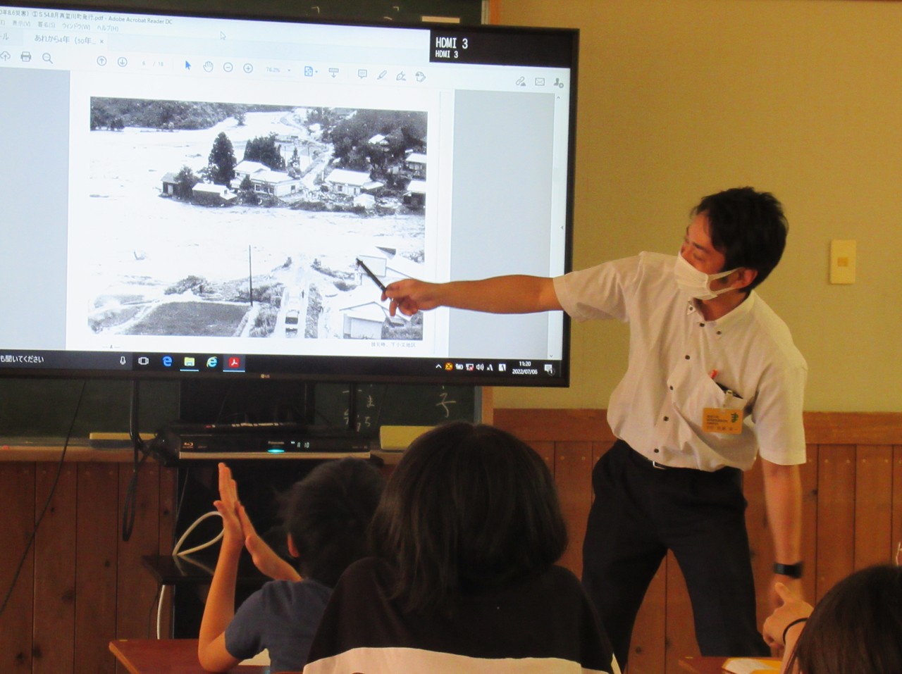 座った小学生の前で胸元にカードを付けた男性がテレビに映った白黒写真をペンで指している写真