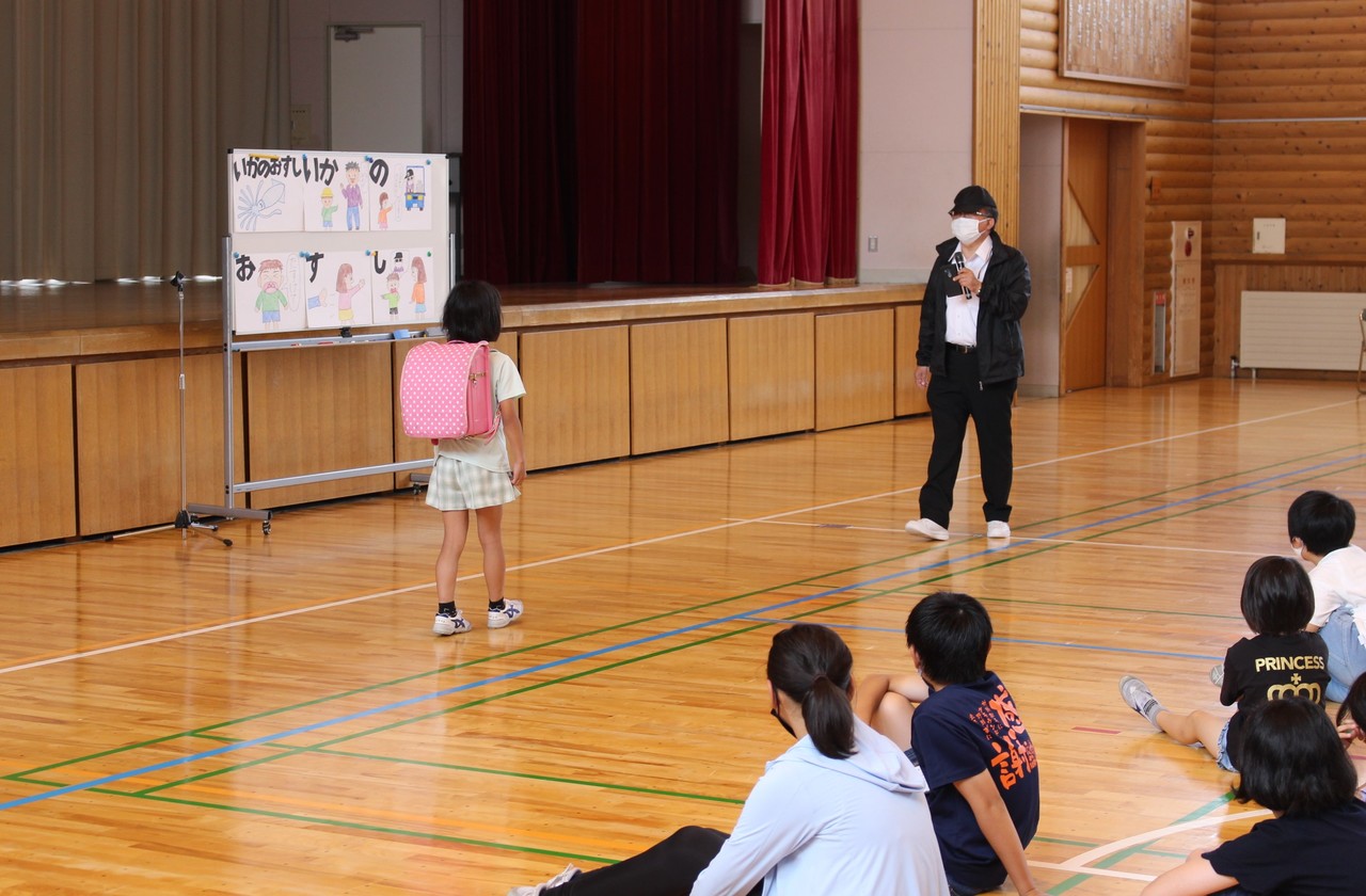 体育館で、ランドセルを背負った女の子と黒い帽子にマスク姿の男性が向き合っているところを座って見ている子供たちの写真