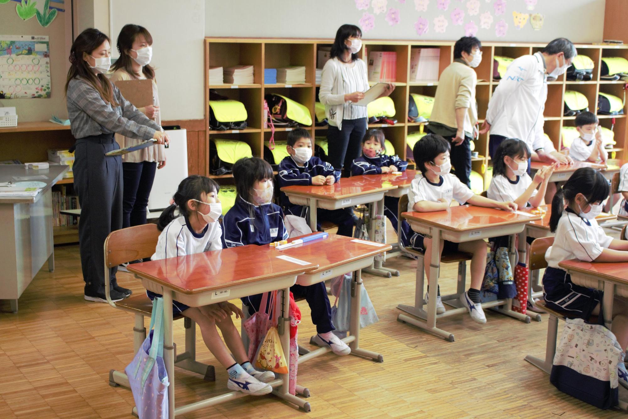 学校の教室の中、着席する体操服姿の小学生と後ろで小学生を眺める5人の男女の写真