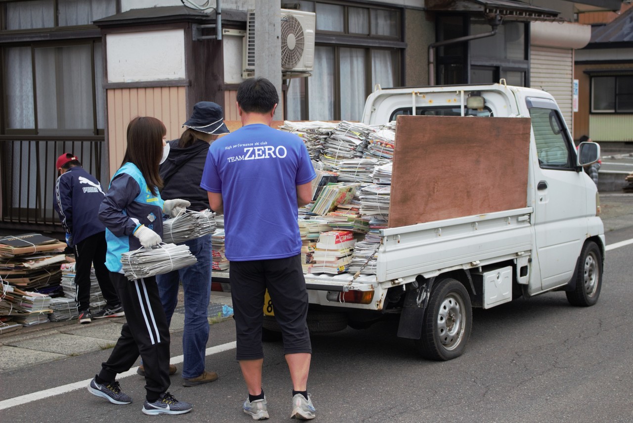 トラックの荷台に古紙が積まれたところに更に古紙を運び入れる人々の写真
