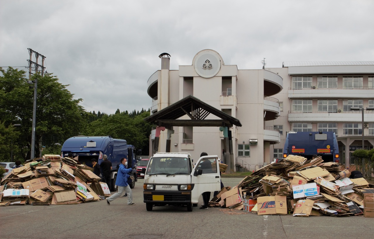 建物の前にトラックが3台停まり、その周りに大量の古紙が山のように置かれた写真