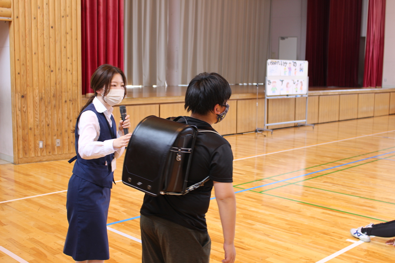 体育館で、制服姿の女性がランドセルを背負った子供にマイクを持って話しかけている写真