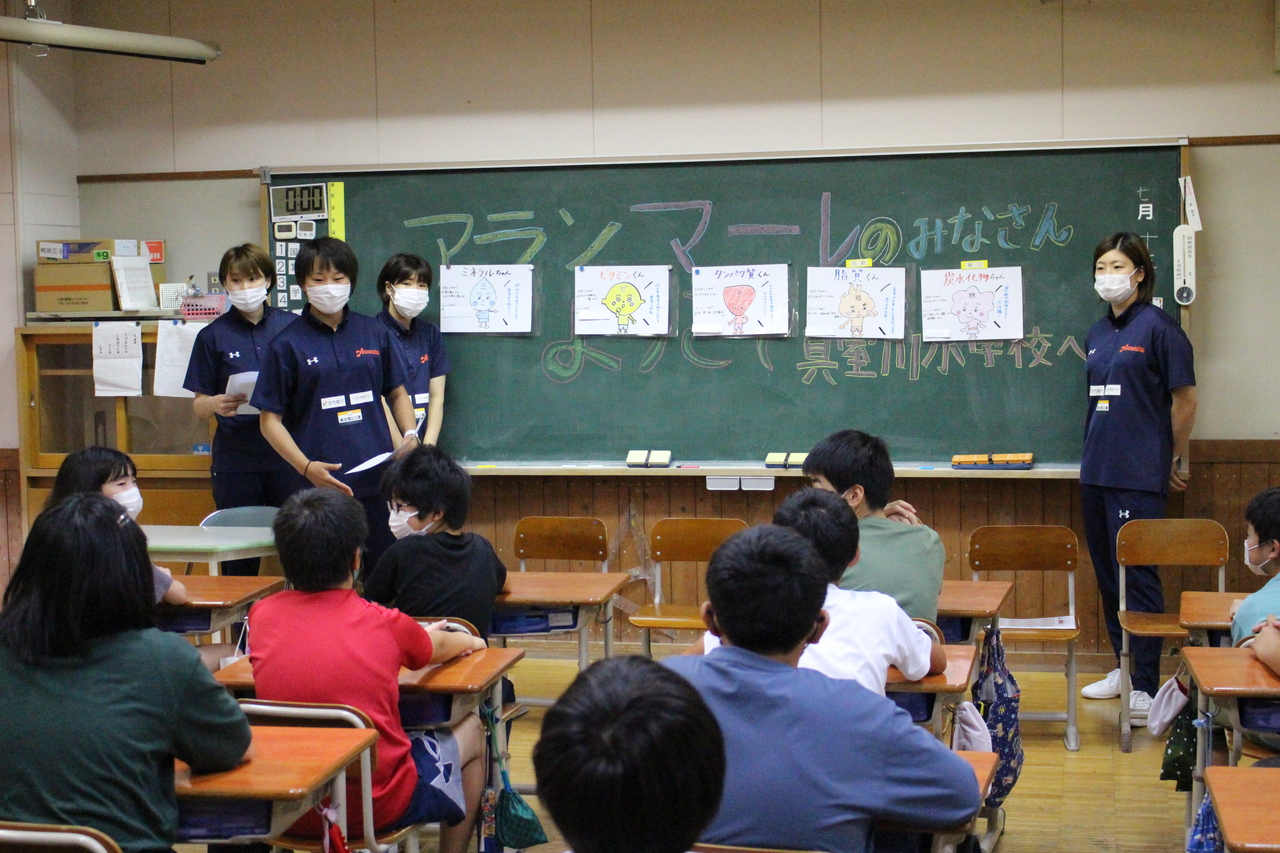 キャラクターの描かれた5つの紙が貼られている黒板の前に立つ4人の女性と、席についている子供たちの写真