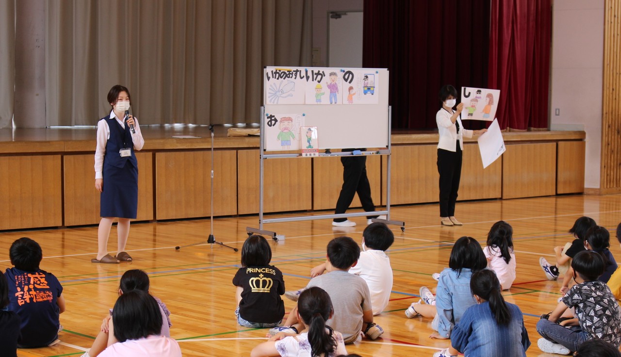 体育館で、子供たちが床に座り、ホワイトボードに貼られた「いかのおすし」と書かれた紙やマイクを持った制服姿の女性を眺めている写真