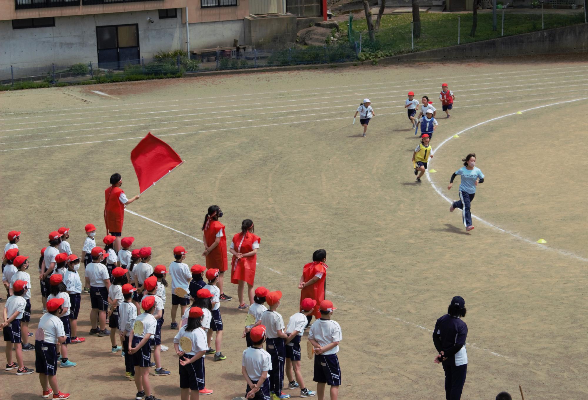 校庭の中、コースを走る子供たちとそれを眺める体操服姿の子供たちと赤色の旗を持った応援団の写真