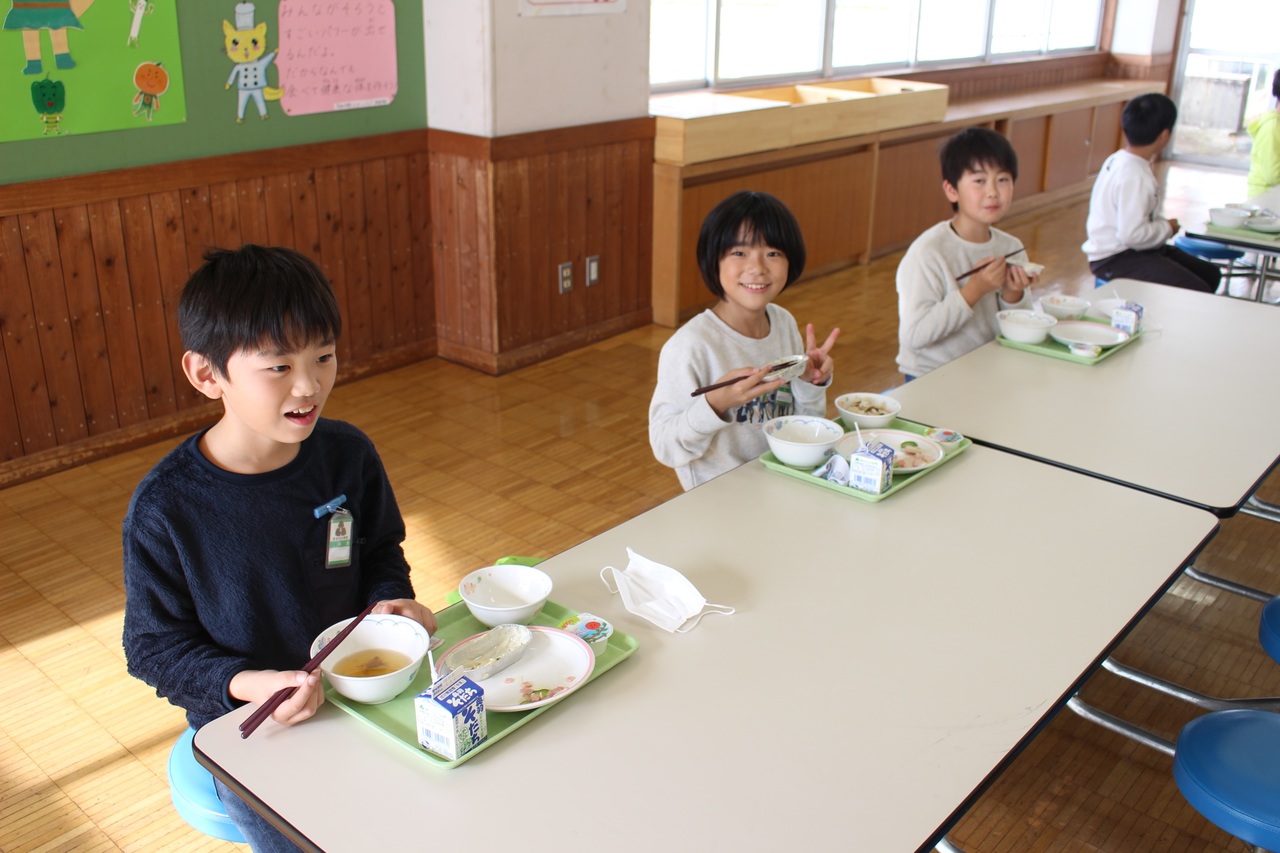 机につき給食を食べて笑顔を見せる子供たちの写真