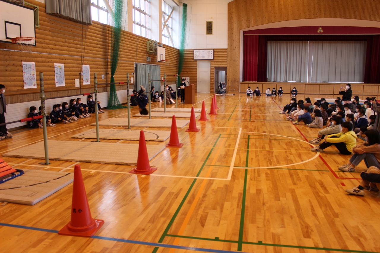 体育館に設置された鉄棒で回転する子供と体育座りをしてそれを見ている子供たちの写真