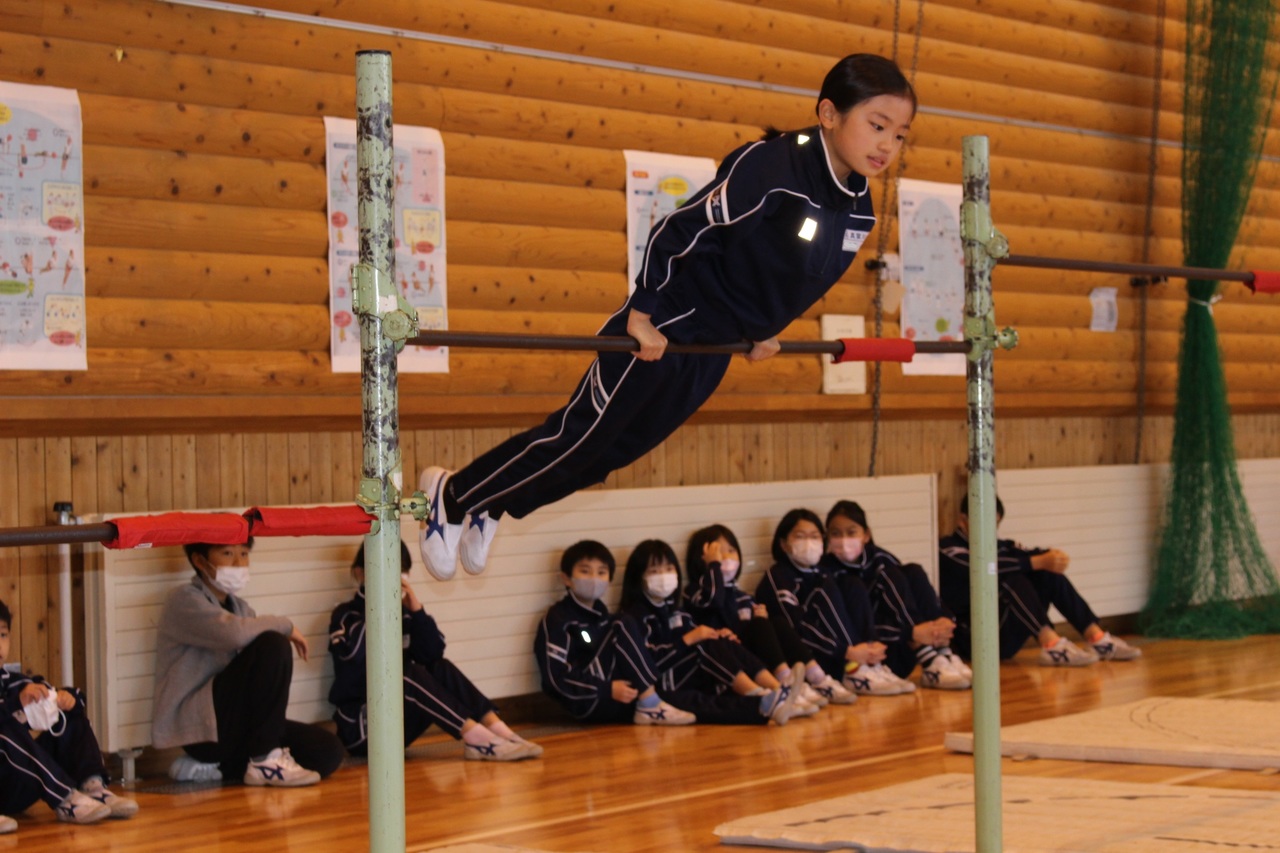 鉄棒に乗ってバランスをとっているジャージ姿の女の子とその後ろで体育座りをしてそれを見ている子供たちの写真