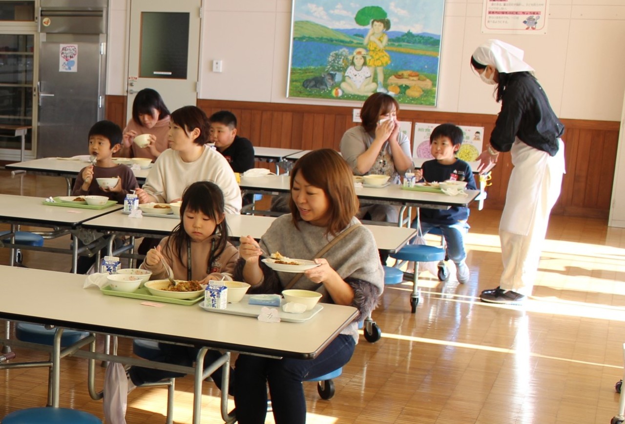 複数の親子が給食を食べている中、一組の親子に声を掛けるエプロン姿の女性の写真