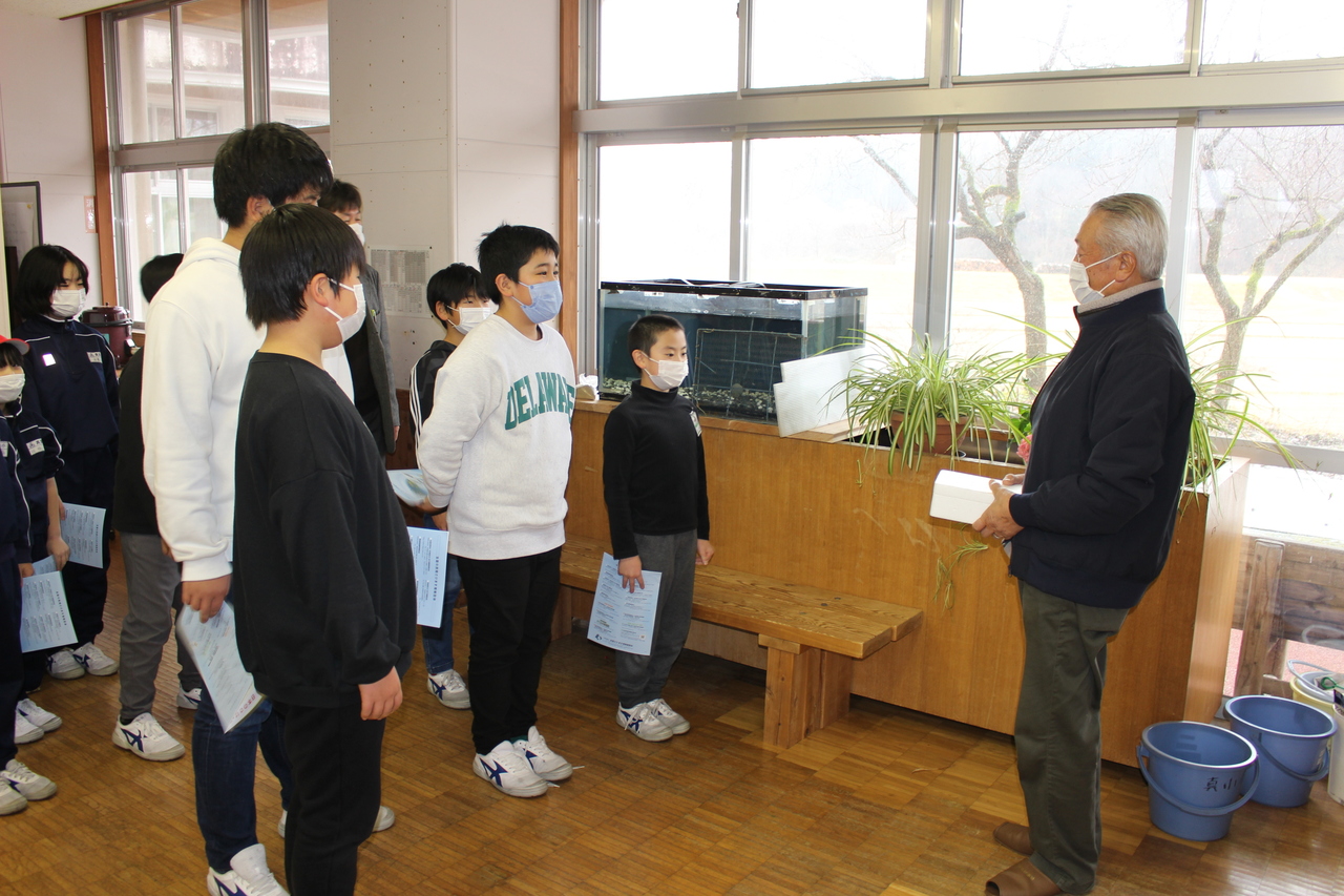 壁際に置かれた水槽の横で、白い箱を持った男性と青いプリントを持った子供たちが向き合っている写真