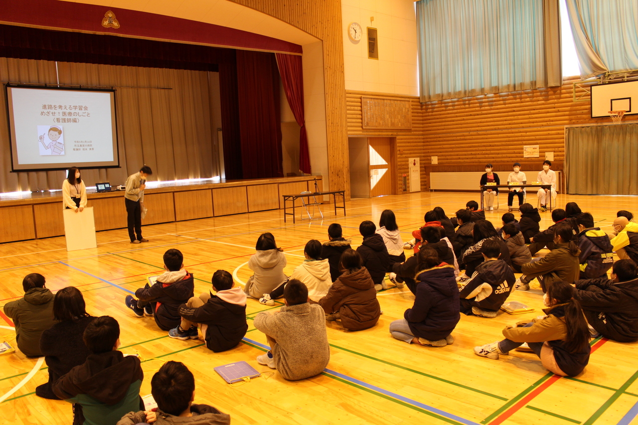体育館に座り男性と女性の話を聞いている児童たちの写真