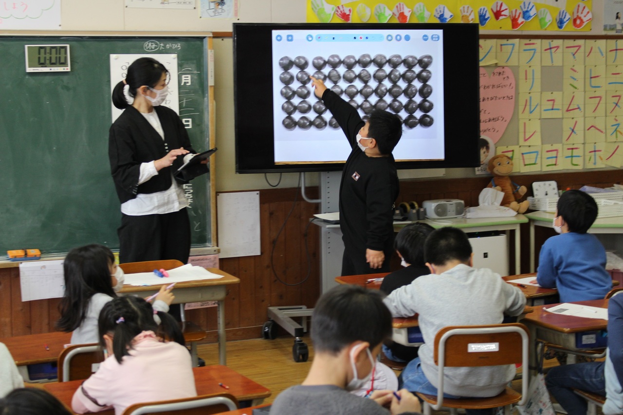 学校の教室で子供たちが席につく中、前方にあるモニターを指さす男の子の写真